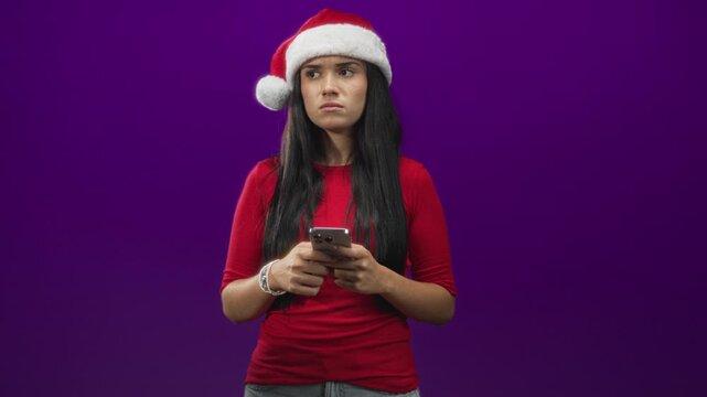 Woman in santa hat holds smartphone with both hands and looks sideways with a frown, against a purple studio backdrop; holiday skepticism.