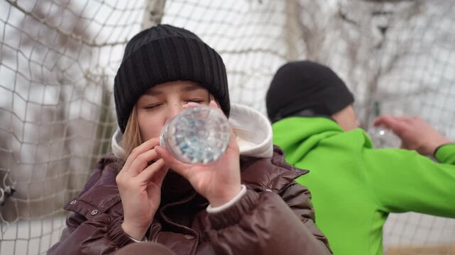caucasian woman with black beanie delicately sipping from bottle cap near goal net, intimate closeup showing concentration and cold breath, reflective outdoor training pause with muted winter tones.