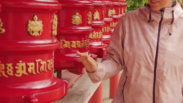 Woman in a pink jacket turns a row of red and gold Buddhist prayer wheels. Peaceful scene of spiritual practice at a temple. Concept for travel, culture, meditation, and mindfulness