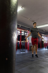 Fototapeta premium Mid-adult man jumping rope in gym near heavy bag, pull-up rig wearing grey t-shirt red shorts