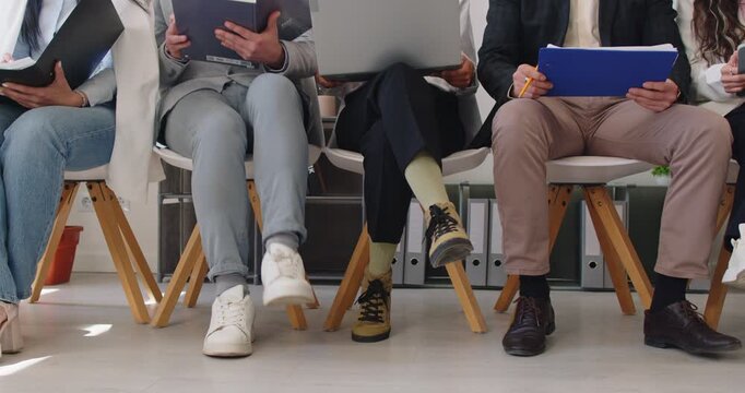 Legs of business people sitting on chairs in a row with resumes and laptops preparing for meeting or conference. Job candidates seekers waiting for interview invitation turn. 4k video.
