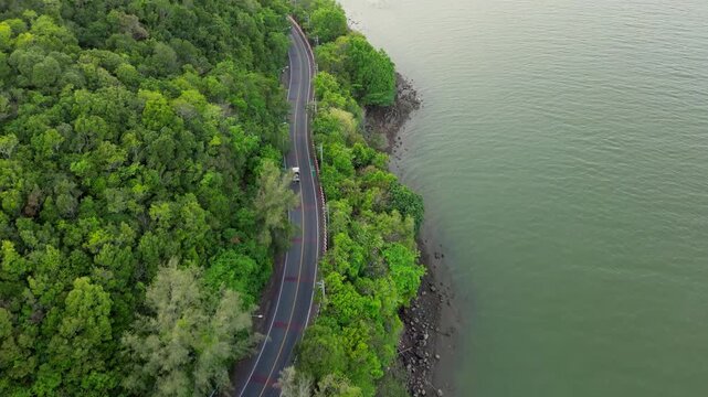 Drone footage reveals lush green forest flanking a winding road along the water. A paved path curves along the tranquil natural edge of the river, creating a beautiful arc.