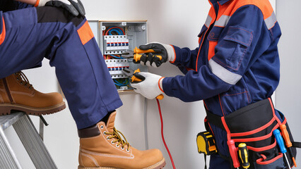Two electricians are working together on an electrical distribution board. ​One technician is sitting on a ladder while the other stands beside the panel. ​safe and professional working environment. © Jakub