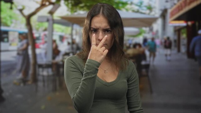 Young hispanic woman points two fingers to nose at street cafe terrace outdoors while seated at a table, wearing a heart pendant; annoyance impatience.