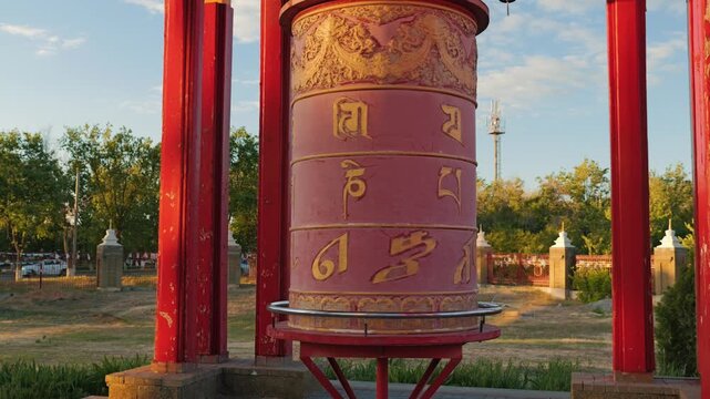A large red Buddhist prayer wheel with golden mantras spins at sunset. This traditional religious object is bathed in the warm light of the sunset, creating a serene and spiritual atmosphere