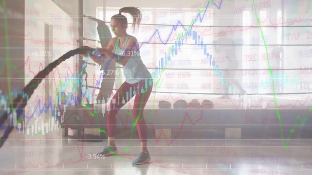 Female athlete gripping ropes and ramping waves for power in gym, stock charts overlaying
