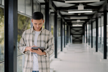 Fototapeta premium Young asian man looking at his tablet in a modern glass hallway. working online. Work from anywhere.