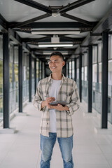 Fototapeta premium Smiling asian man standing and look at to the window holding tablet in a modern glass hallway. working online. Work from anywhere. modern tech