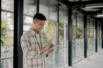 Fototapeta premium Young asian man smiles while writing on tablet beside large glass window. Working outdoor. Chilling Outside, Urban life. 