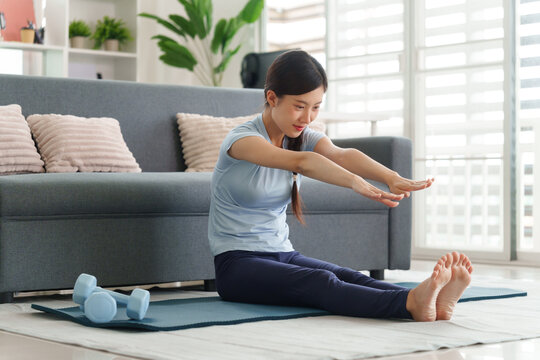 Asian adult woman stretching doing home yoga workout on floor mat with dumbbells in modern apartment living room practicing healthy fitness routine