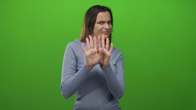 Woman with hands raised showing palms and grimace, recoil gesture, wearing blue sweater in studio against vivid green backdrop; disgust.