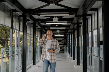 Obraz premium Close-up happy young Asian man walking, looking outside and talking phone with client and holding tablet on his hand in a modern glass hallway. Work from anywhere. Remote work. outdoor.