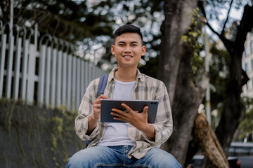 Fototapeta premium Handsome asian man student or office worker holding tablet while sitting on concrete chair with urban city on the background. Working or Study, Modern Tech, Chilling Outdoors. Remote work.