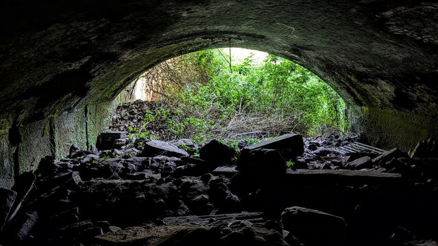 Inside abandoned tunnel, curved arched walls. Dark stones, collapsed underground passage. Daylight cave contrast light at the end of the tunnel, bushes greenery. Rocks, shadows, under the bridge arch.