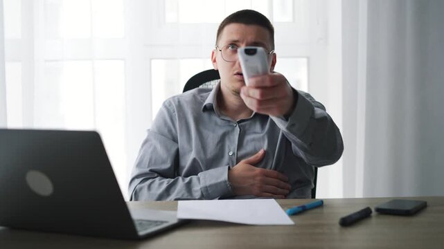 A businessman turns on the air conditioner in his office to warm up.
The office is freezing, and a man is shivering with the air conditioner remote control.