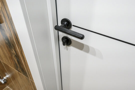 A sleek white interior door with a matte black lever handle and matching lock. Set against a wood-patterned tiled wall, featuring clean lines and a modern minimalist design.