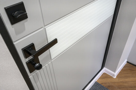 A modern white intercom door with a sleek black handle and matching lock, featuring frosted glass panels and textured detailing. Set against a light wall with wooden flooring.