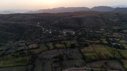 Fototapeta premium Aerial view of the Aravalli hills near Udaipur, Rajasthan at sunset, showing winding roads, patchwork agricultural fields, native trees, and scattered village houses across rugged terrain.