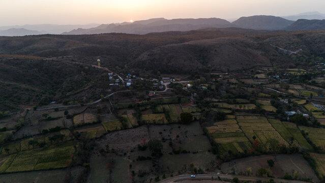 Aerial view of the Aravalli hills near Udaipur, Rajasthan at sunset, showing winding roads, patchwork agricultural fields, native trees, and scattered village houses across rugged terrain.