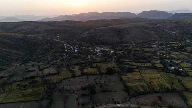 Aerial view of the Aravalli hills near Udaipur, Rajasthan at sunset, showing winding roads, patchwork agricultural fields, native trees, and scattered village houses across rugged terrain.