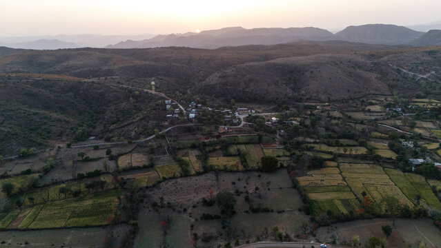 Aerial view of the Aravalli hills near Udaipur, Rajasthan at sunset, showing winding roads, patchwork agricultural fields, native trees, and scattered village houses across rugged terrain.