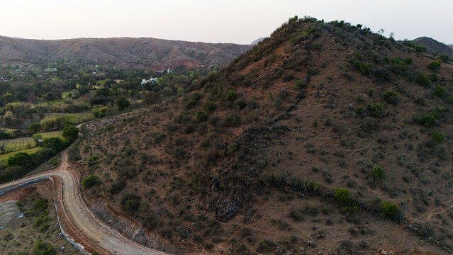 Aerial view of the Aravalli hills near Udaipur, Rajasthan at sunset, showing winding roads, patchwork agricultural fields, native trees, and scattered village houses across rugged terrain.