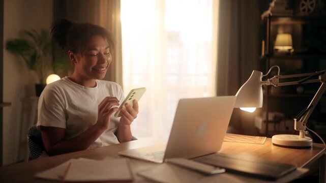 Happy african american woman scrolling on smartphone while sitting at desk with laptop in sunny room
