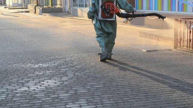 Man in airtight suit sprays disinfecting pavement on street in city. Sanitation workers clean crosswalk. Sanitary measures. Coronavirus pandemic Knapsack sprayer