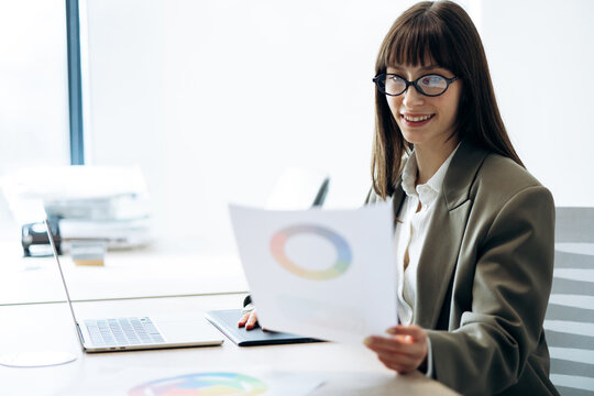 Businesswoman reviewing financial data and marketing reports, using a color wheel chart while working on a laptop at her office desk