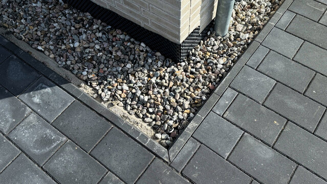 Close-up view of a corner of a paved patio featuring gray interlocking stones and decorative gravel surrounding a brick column with a drainage pipe