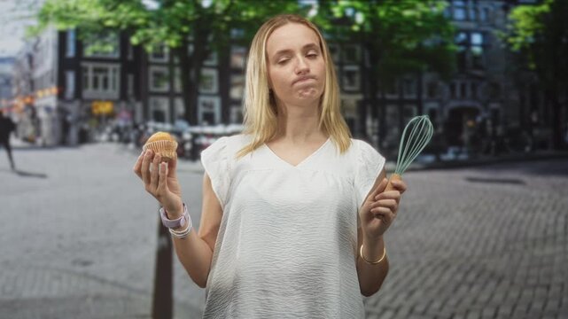 Woman wearing white blouse holding muffin and whisk with a resigned expression on a cobblestone street in a european canal district; culinary disappointment.