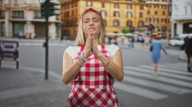 Woman chef in red gingham apron presses palms together in pleading gesture at a busy street crosswalk with blurred pedestrians and cars; hopeful.