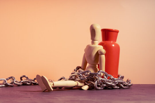 Wooden mannequin chained to red vase while seated against it, confinement and dependency concept