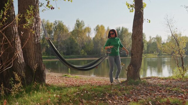 Caucasian woman setting hammock by lake, attaching straps to trees, smoothing fabric and testing sag autumn foliage, calm water reflection, soft sunlight, solo camper preparing quiet campsite