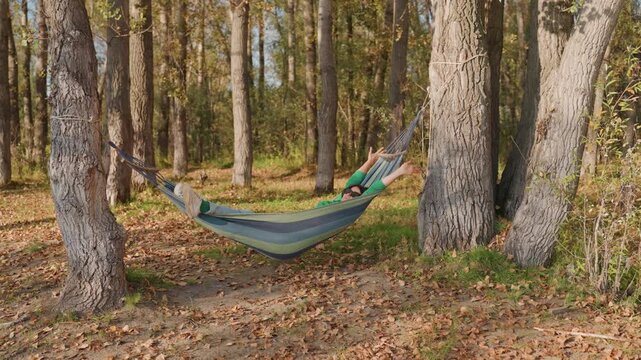 White male camper napping in hammock under birch canopy golden autumn light warms textured bark and leafstrewn floor gentle breeze rocks hammock during outdoor travel break, evoking restful travel