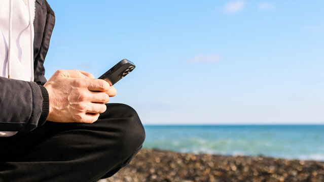 Person in hooded jacket using phone on pebbly beach