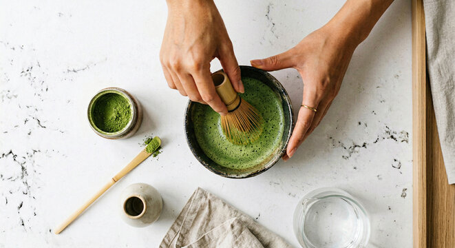 Hands whisking matcha green tea powder in ceramic bowl overhead view