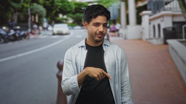 Young hispanic man points finger down at brick sidewalk on a street beside a bollard, wearing unbuttoned shirt and black tshirt; playful confidence.