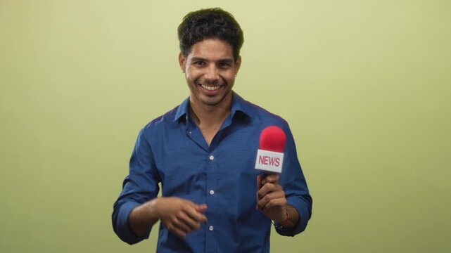 Young hispanic man smiling and pointing finger at red news microphone held in studio with plain green backdrop; confidence upbeat.