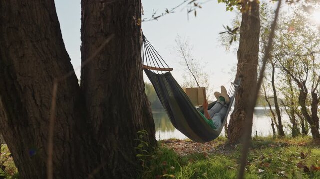 Hammock by lake camping person reading book, shaded grove of trees, sunlit water reflections, gentle breeze, grassy shore, calm afternoon, unplugged nature retreat for quiet restoration