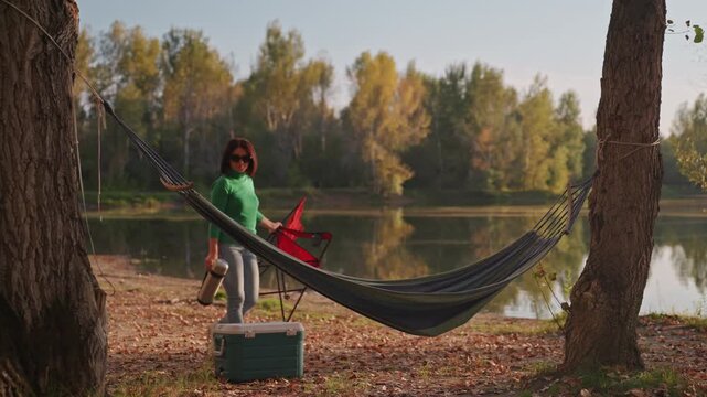 White woman camping hammock relaxes lakeside, sips mug, watches water, gentle breeze through trees, cooler and chair beside hammock, golden dusk light, tranquil solitude and slow unwind outdoors.