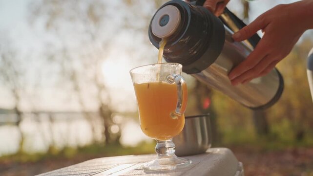 Lakeside camping morning orange juice pour from stainless thermos into glass goblet with sugared rim, camper hand holding thermos, warm golden sunlight, calm lake reflection, soft bokeh background,