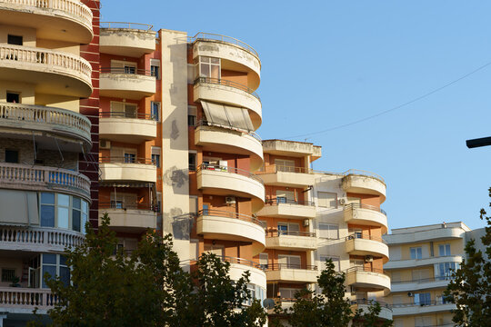 Sunlit facade of aged residential buildings with curved balconies and peeling paint. Terracotta and cream colors contrast with a clear blue sky, showing weathered urban architecture.