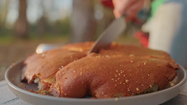 Hands cutting skillet cornbread at campsite, closeup of crumbly crust breaking into pan, vigorous slices by camper hand, crumbs scattering on metal rim, trees and gear blurred in background conveying