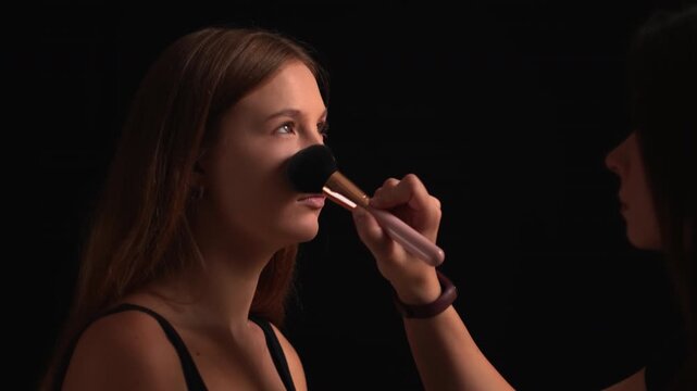 Professional makeup artist applying foundation with a brush on a beautiful young woman's face during a backstage makeover session in a dark studio with dramatic lighting