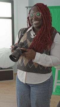 Woman holds camera and smiles while adjusting the lens in a bright photo studio, wearing round glasses and an argyle sweater vest; creative friendly joy.