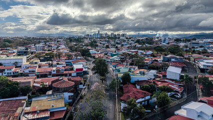 Obraz premium Drone aerial view of downtown San Jose, Costa Rica, showcasing dense urban neighborhoods, city skyline, dramatic cloudy sky, a vibrant Central American capital with mixed architecture (February 2026)