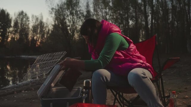 Dusk lakeside camping woman cleaning gear beside folding chair, rinsing fishing net over portable stove and cooler, pink vest and green sweater, tree silhouettes at sunset, lantern glow casting warm