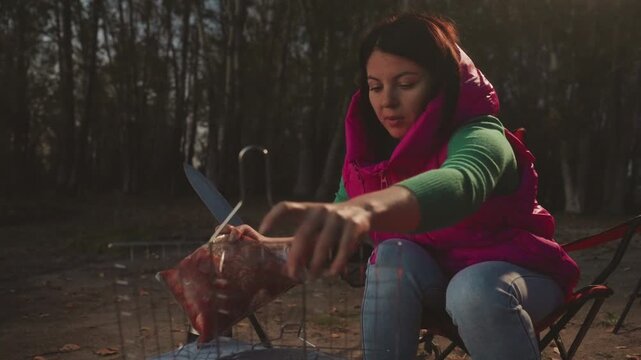 Dusk campsite meat preparation by caucasian woman seated in red folding chair holding knife and raw meat, placing carcass into grill basket, focused camp cook checking marinade and seasoning, campfire