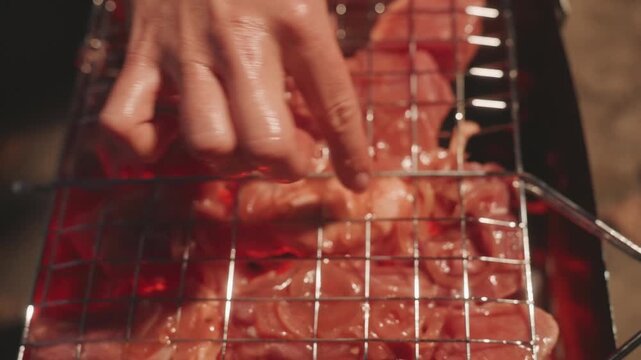 Closeup hand pressing raw meat on metal grate over glowing charcoal at night, tactile marinade and juices glisten, fingers adjust skewer, warm ember light casts red highlights, smoky rustic camping